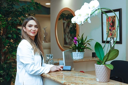 Dentist in a white coat standing behind a reception desk with a computer, smiling, with potted plants and decorative mirror in the background.