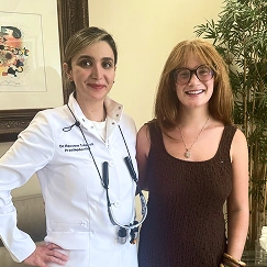 Dentist in a white coat standing next to a patient wearing a dark dress, both smiling at the camera in a dental office.